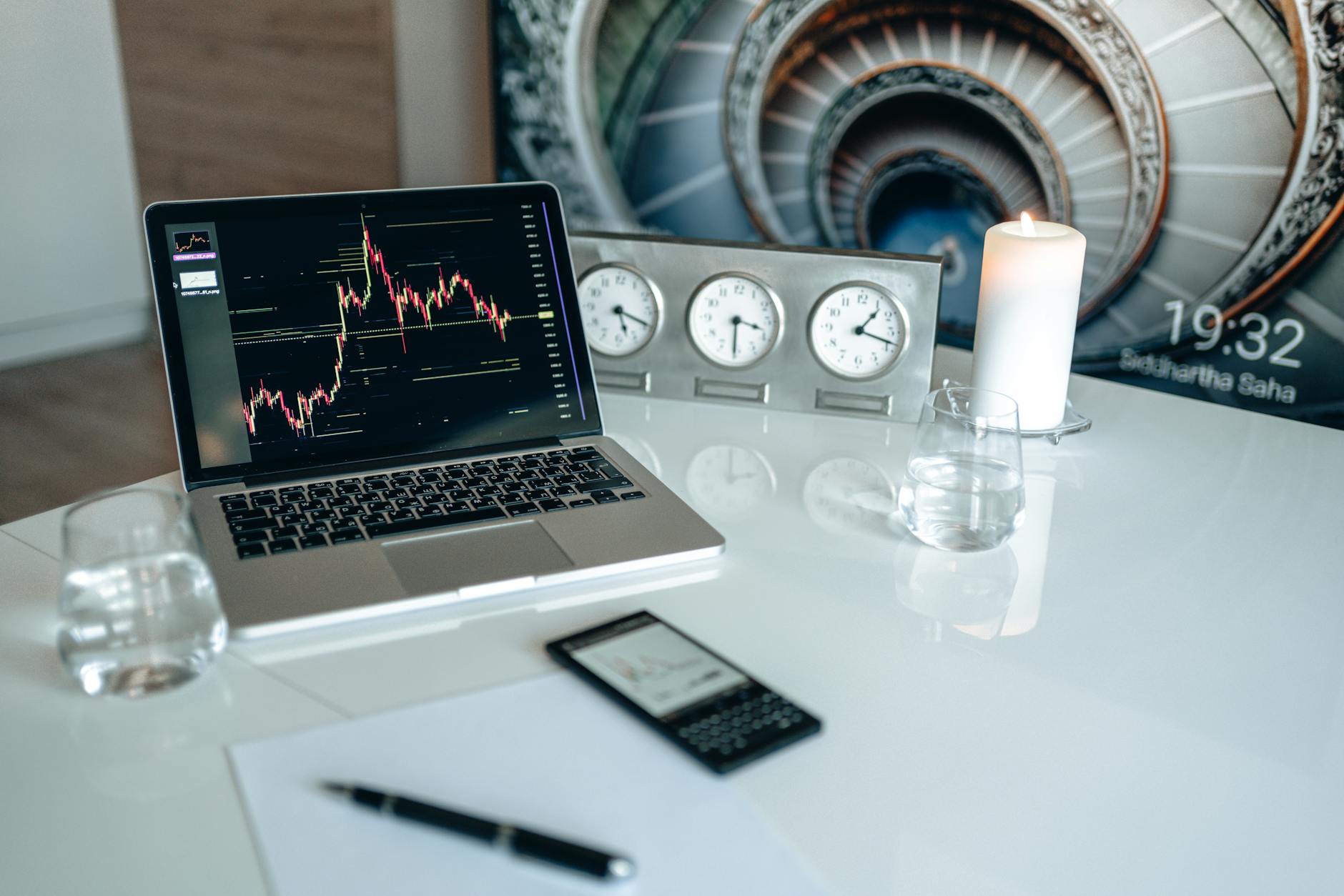 Laptop on desk with trading chart, clocks, candle, glass of water and phone.