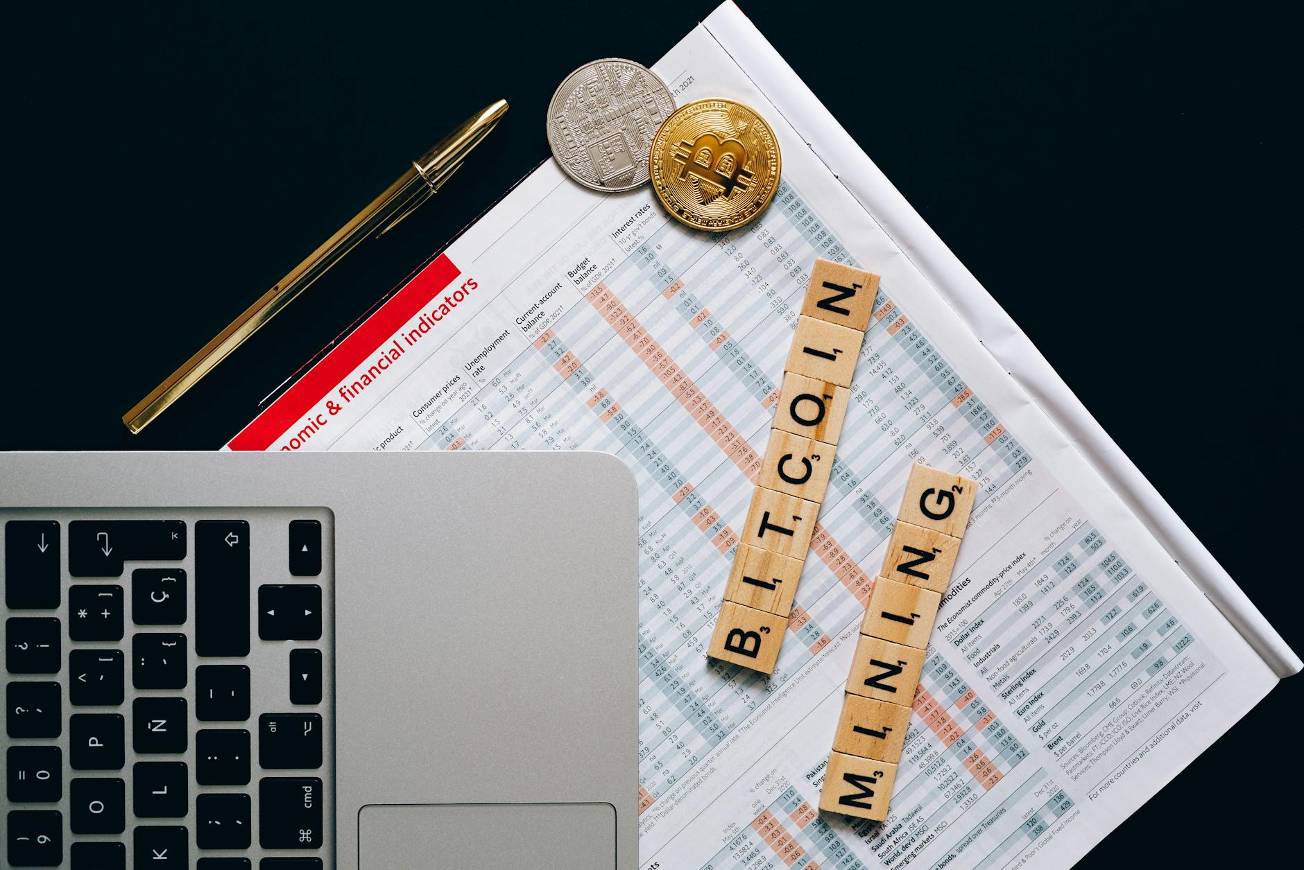 Laptop, pen, Bitcoin coins, and newspaper with economic indicators.