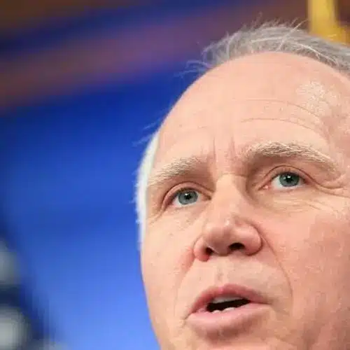 Close-up of a man speaking, with a blue background and American flags behind him.