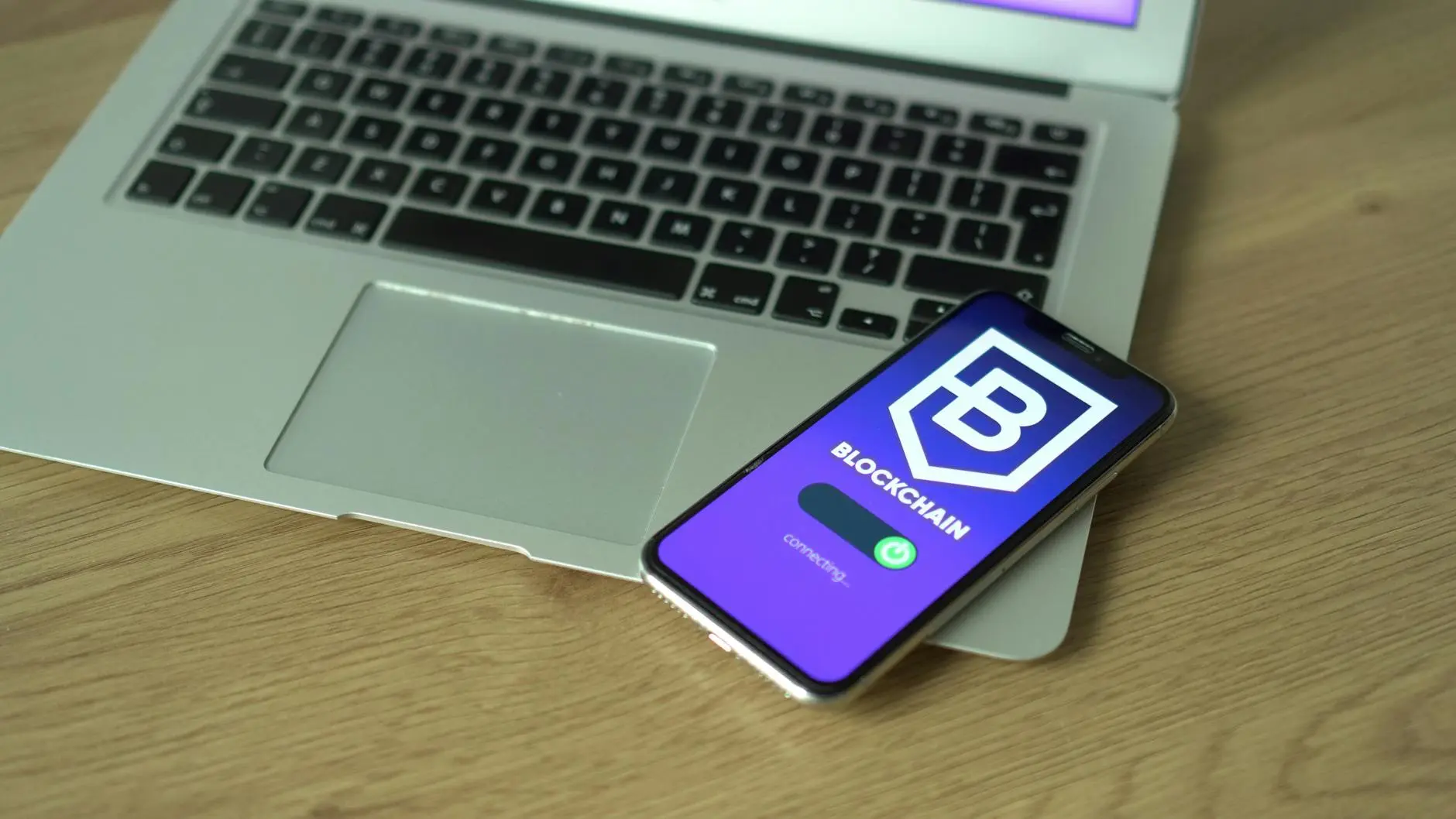 A smartphone displaying a blockchain app interface next to a laptop on a wooden desk.