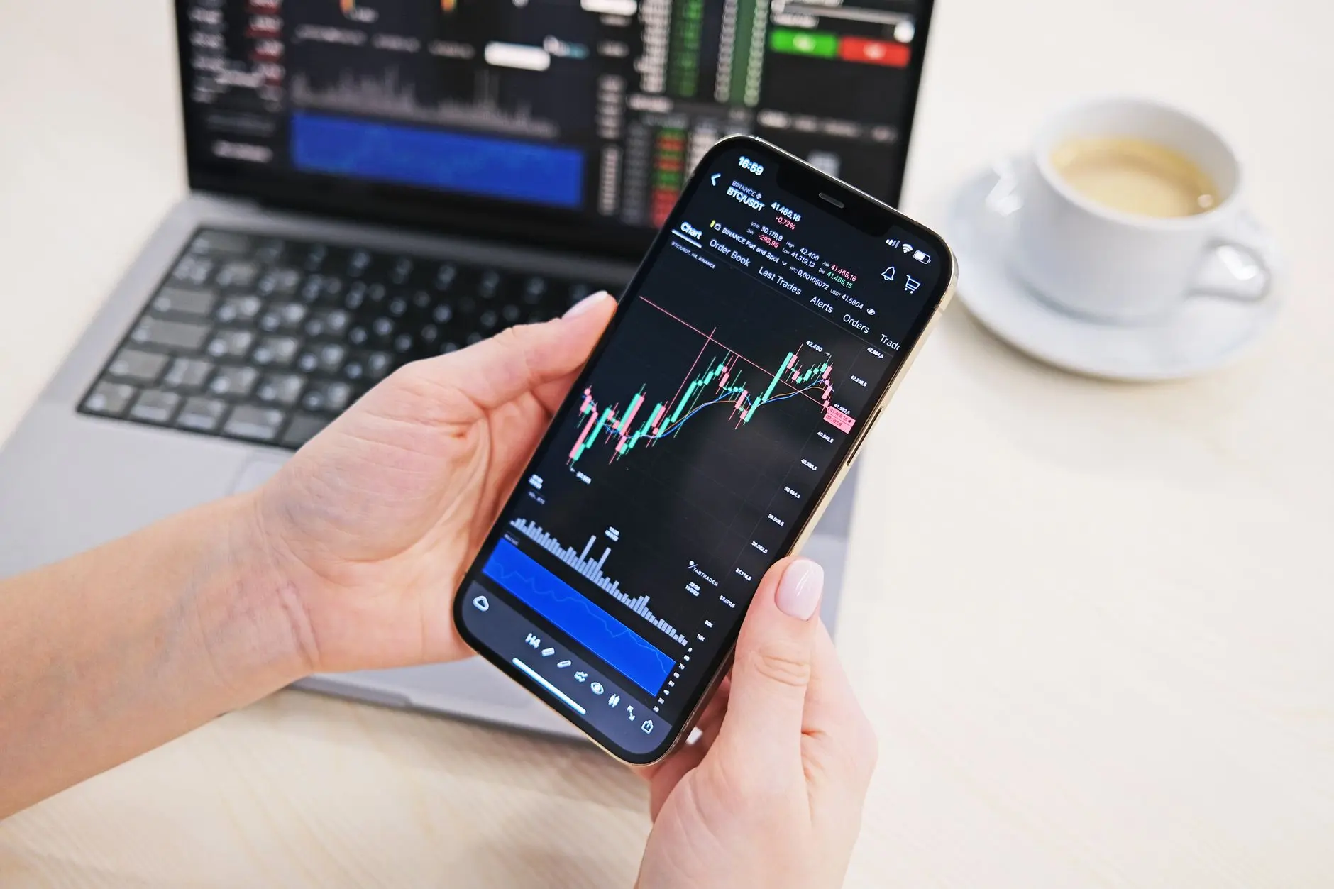 Person holding a smartphone displaying cryptocurrency trading chart with a laptop in background.