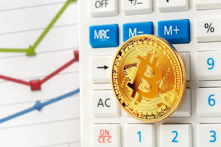 A gold Bitcoin coin resting on the buttons of a white digital calculator next to a financial line graph