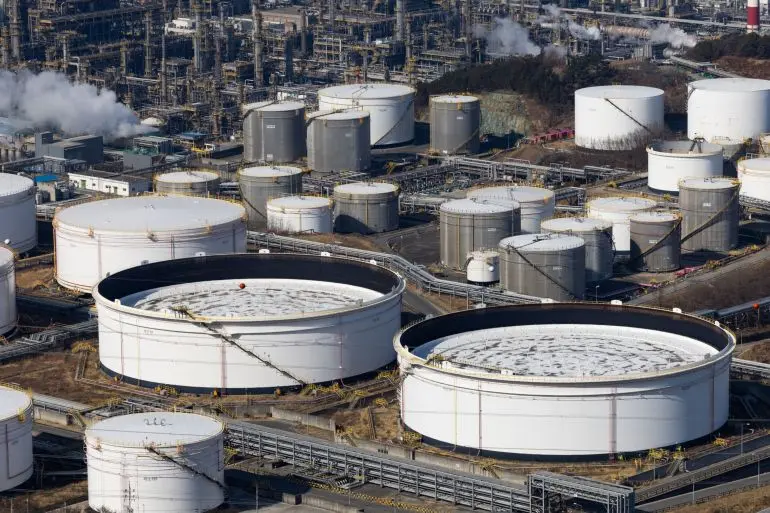 Aerial view of a large oil refinery with numerous white and grey cylindrical storage tanks and industrial piping.