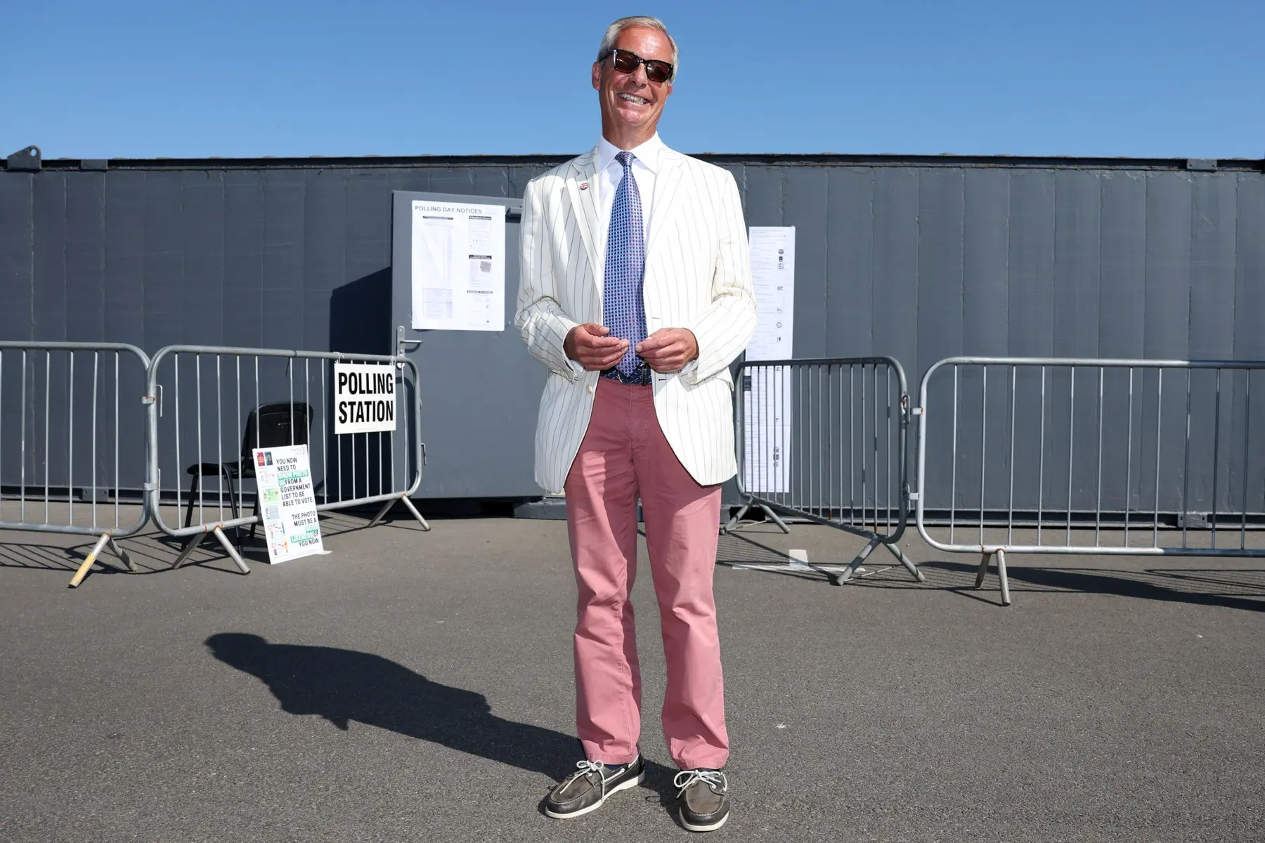 Nigel Farage wearing a white striped blazer and pink trousers standing in front of a polling station.