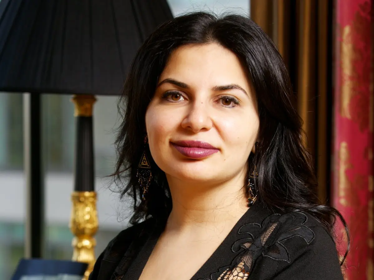 Portrait of Ruja Ignatova with dark hair and purple lipstick, wearing black clothing and ornate earrings.