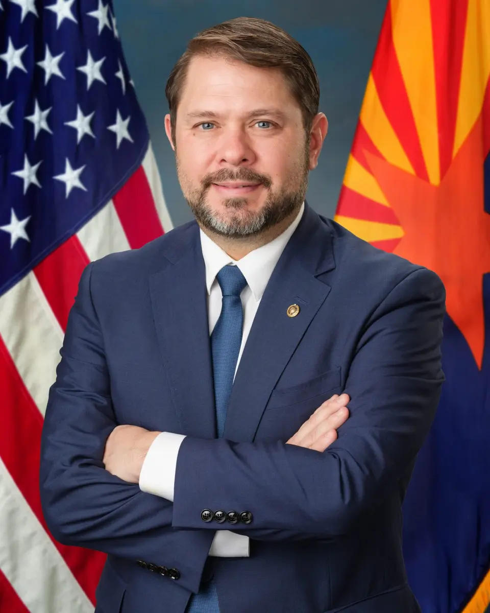 Official portrait of Senator Ruben Gallego in a blue suit, posing with the US and Arizona flags.