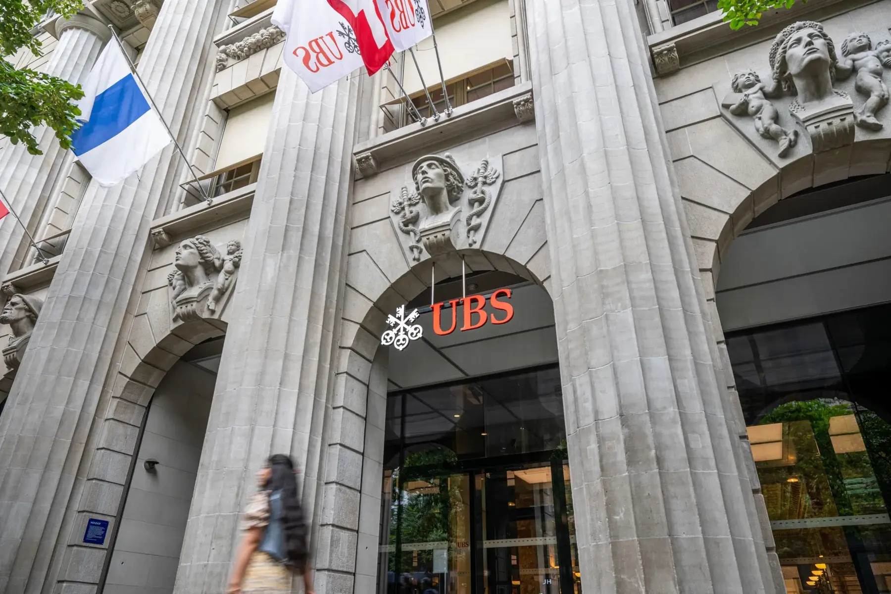 The ornate stone entrance of the UBS Group AG headquarters in Zurich featuring the UBS logo and red flags.