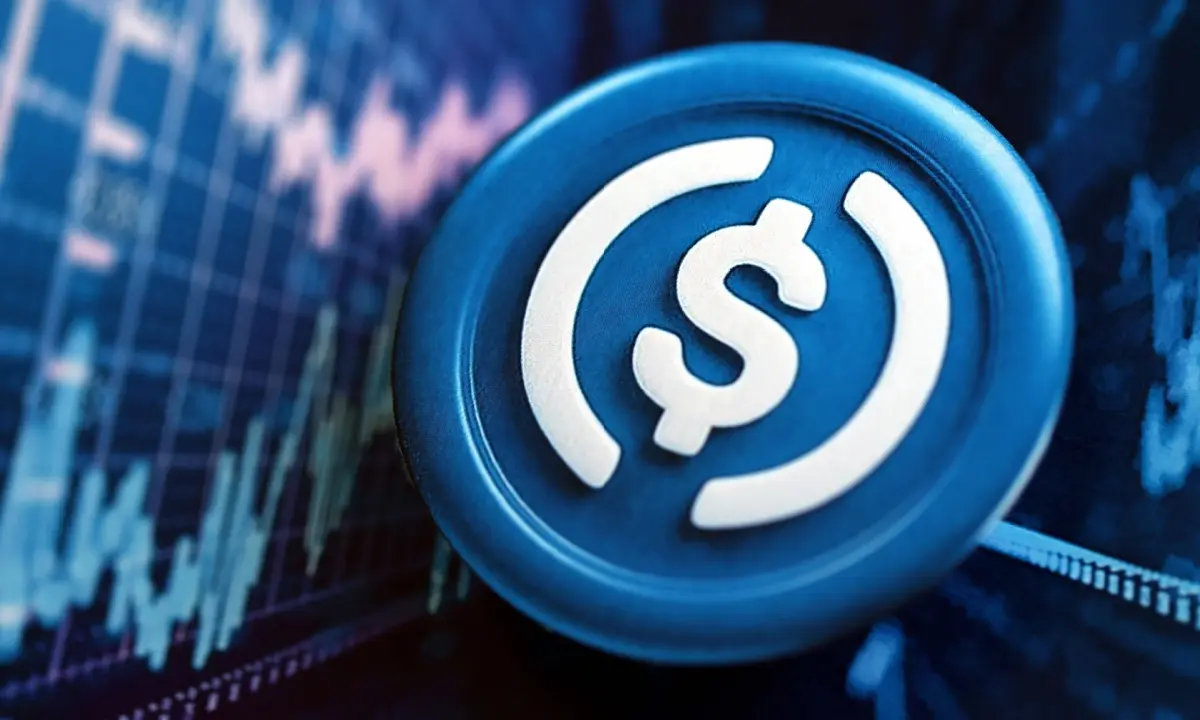 A blue physical token featuring the USDC dollar sign logo against a backdrop of financial charts