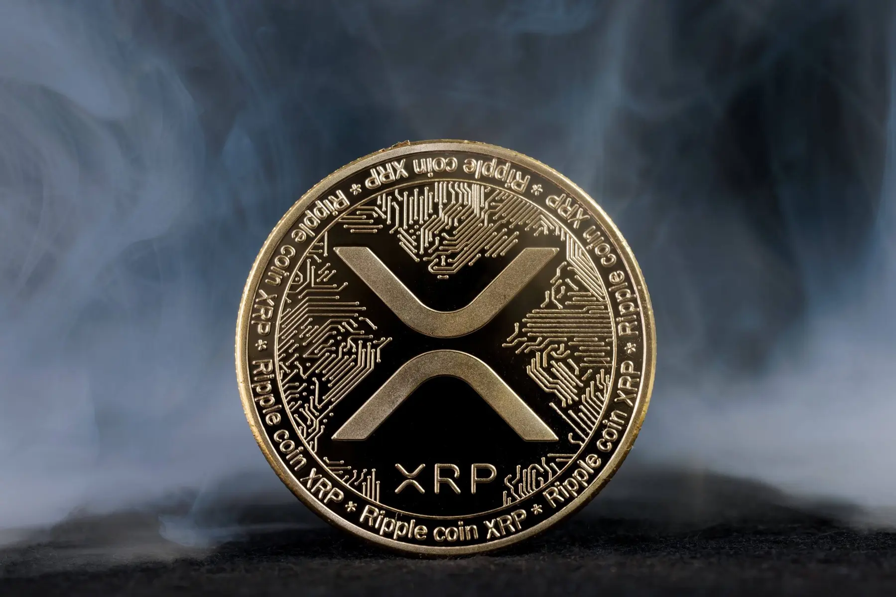 A gold-colored physical XRP Ripple coin featuring the XRP logo and circuit patterns against a smoky background
