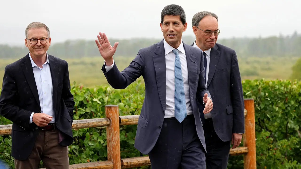 Kevin Warsh in a blue suit and tie gesturing with his hand while walking outdoors with two other men.