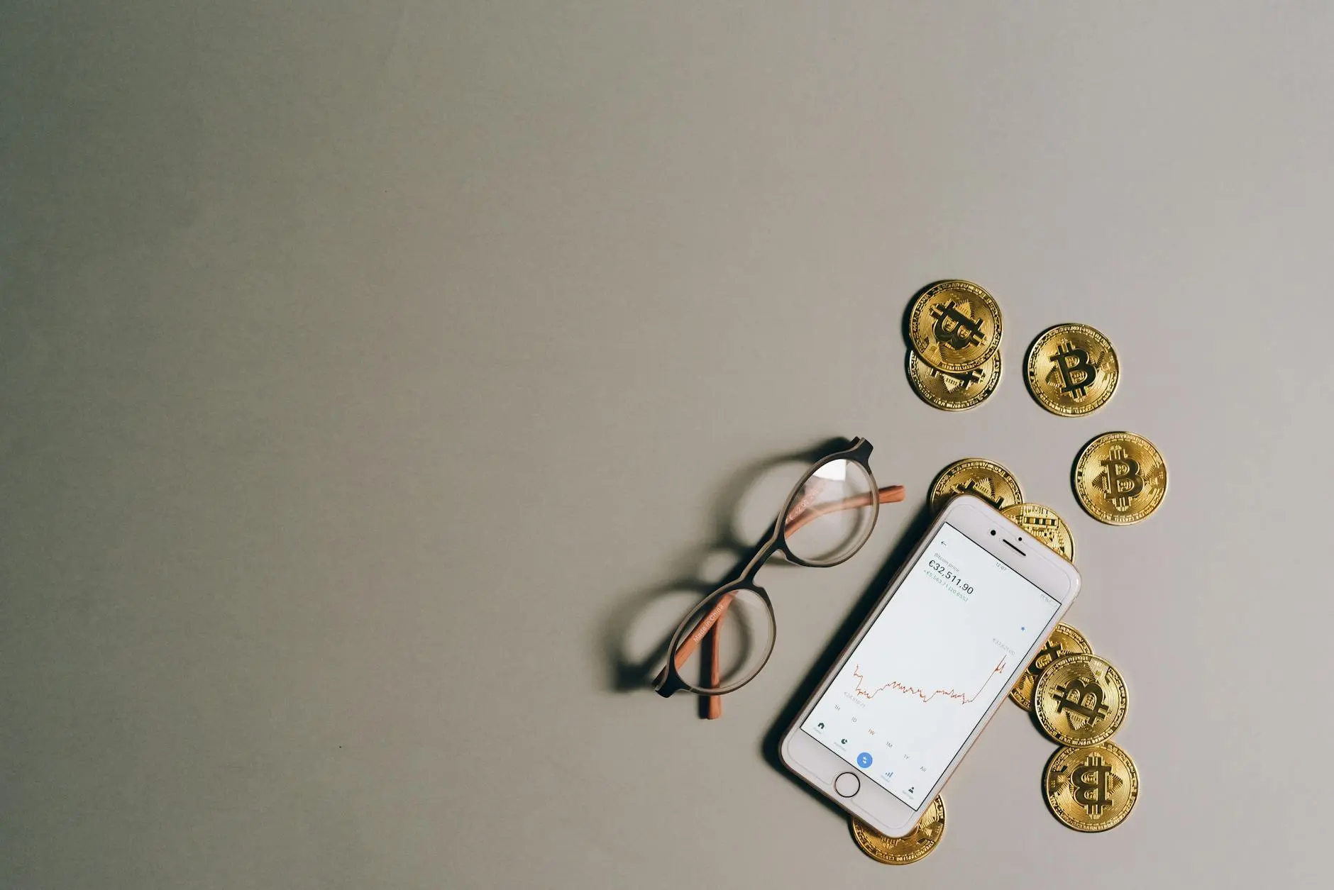 A smartphone displaying a Bitcoin price chart beside gold Bitcoin coins and glasses.