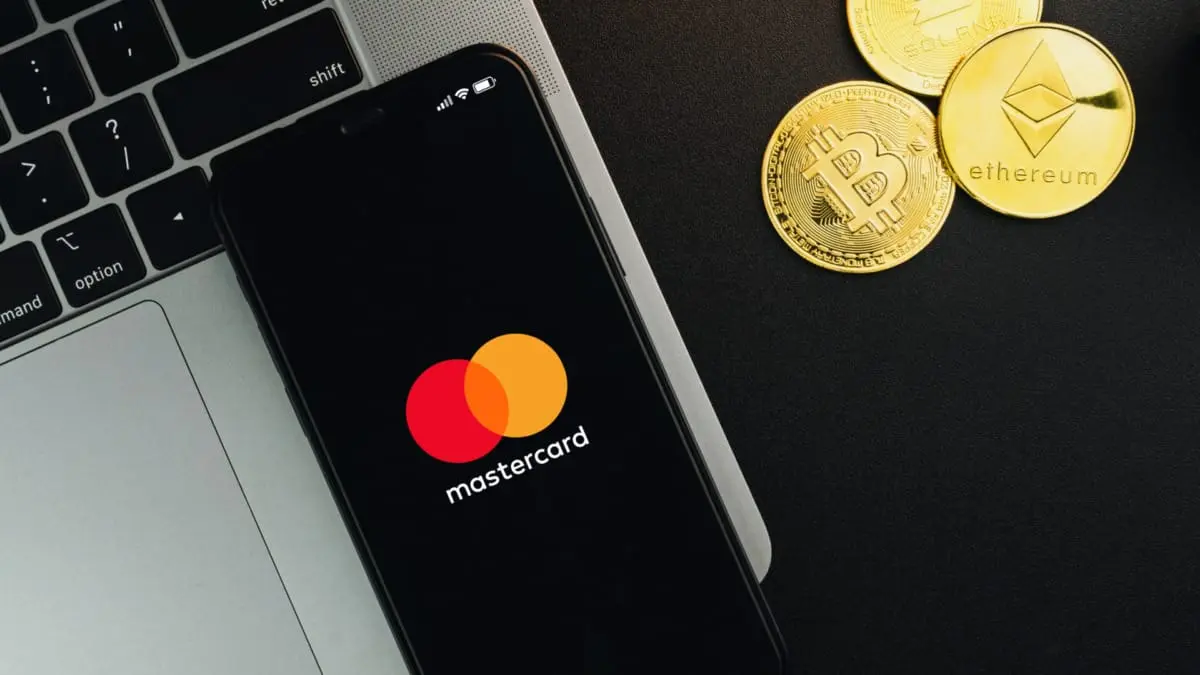 Mastercard logo on a phone beside gold cryptocurrency coins on a laptop.