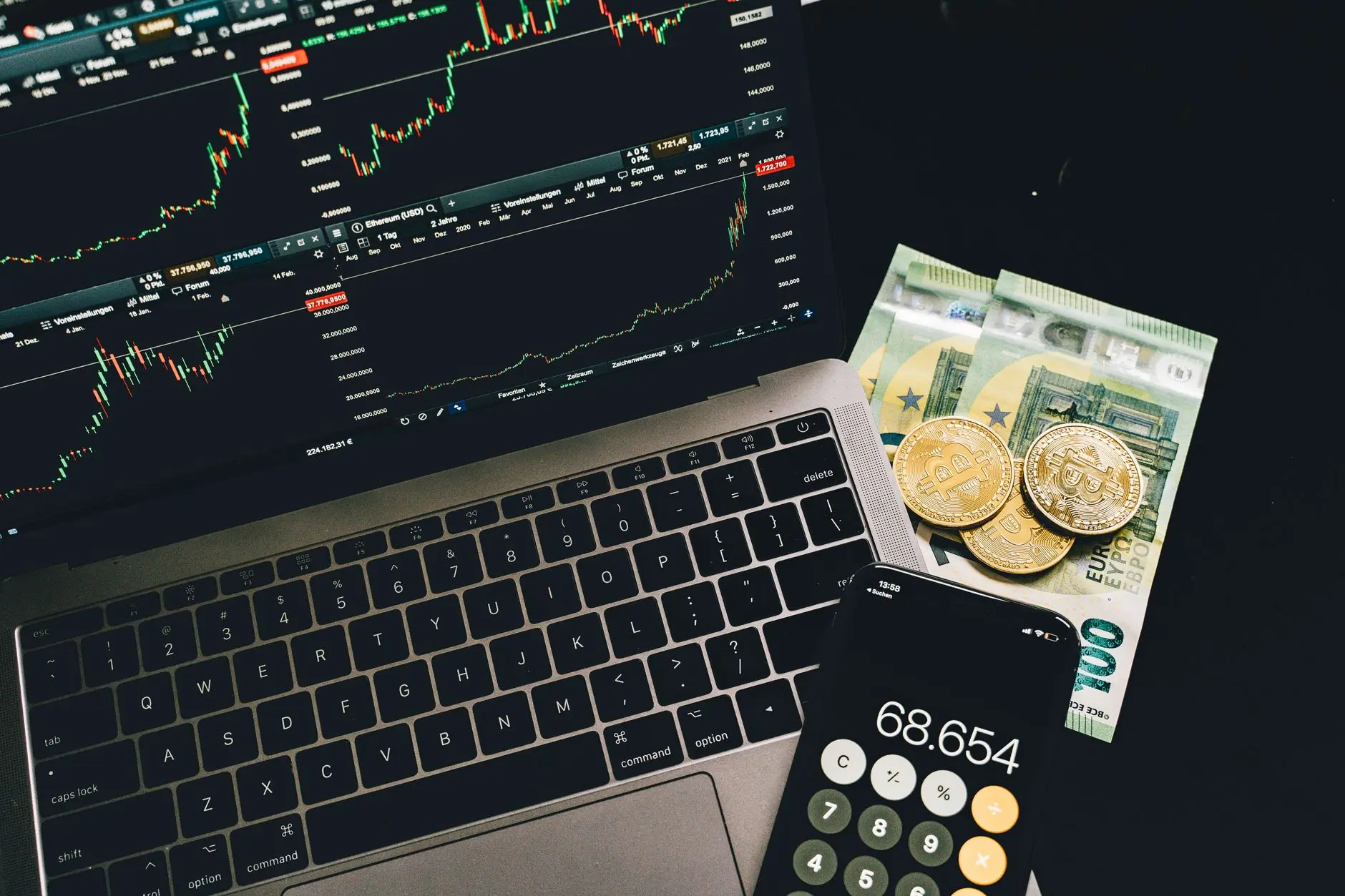 A laptop showing cryptocurrency charts beside euros and Bitcoin coins.