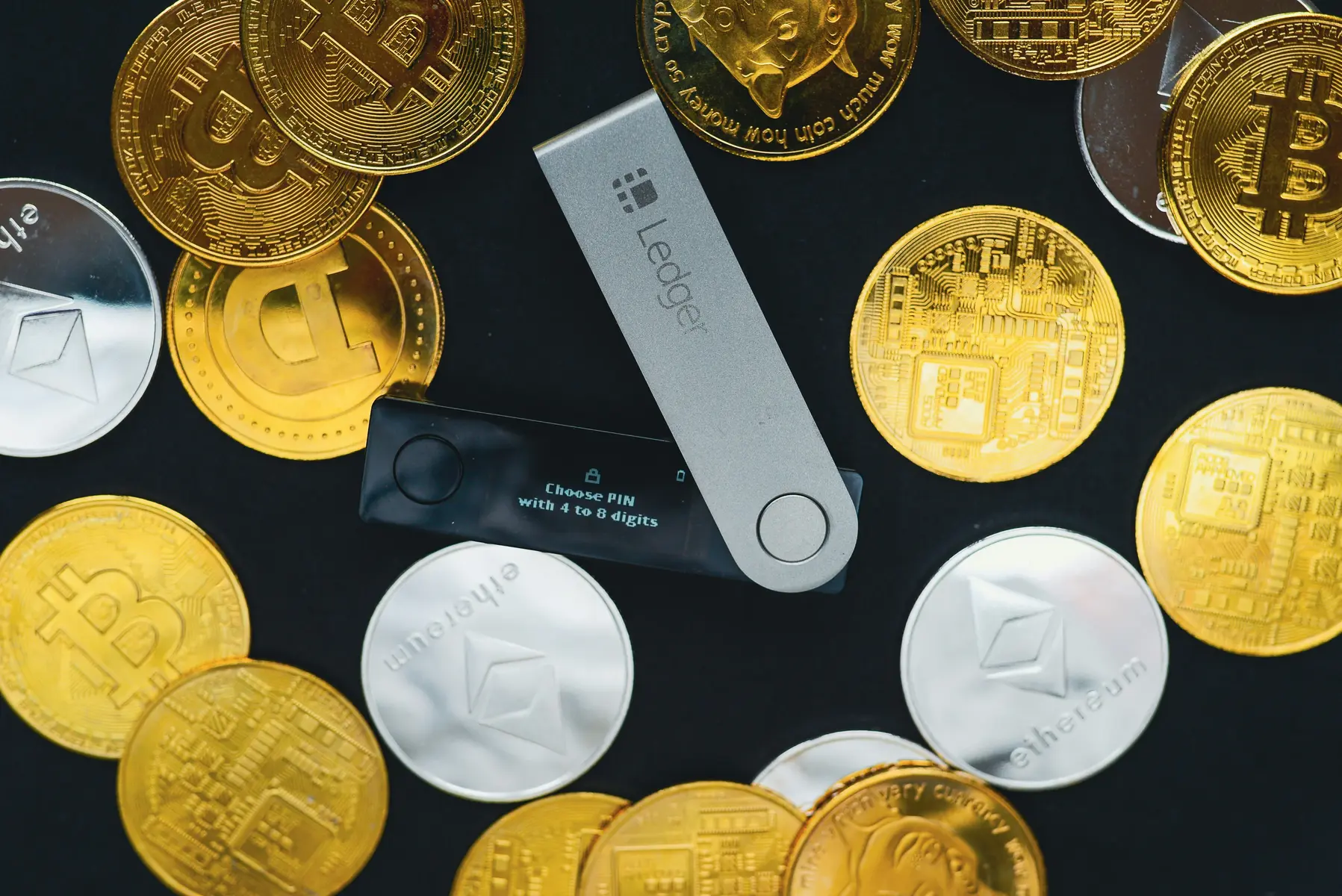 Een Ledger hardware wallet omringd door fysieke Bitcoin- en Ethereum-munten op een zwarte achtergrond.