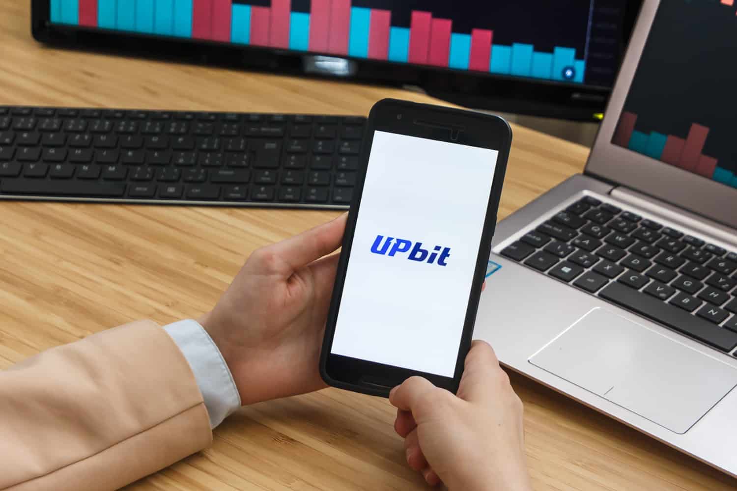 A person holds a smartphone next to two laptop screens. The smartphone displays the Upbit logo.