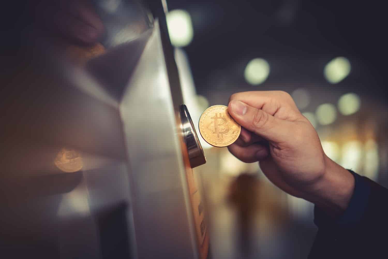 A person’s hand holds a metal coin intended to represent Bitcoin next to a machine with a coin slot.