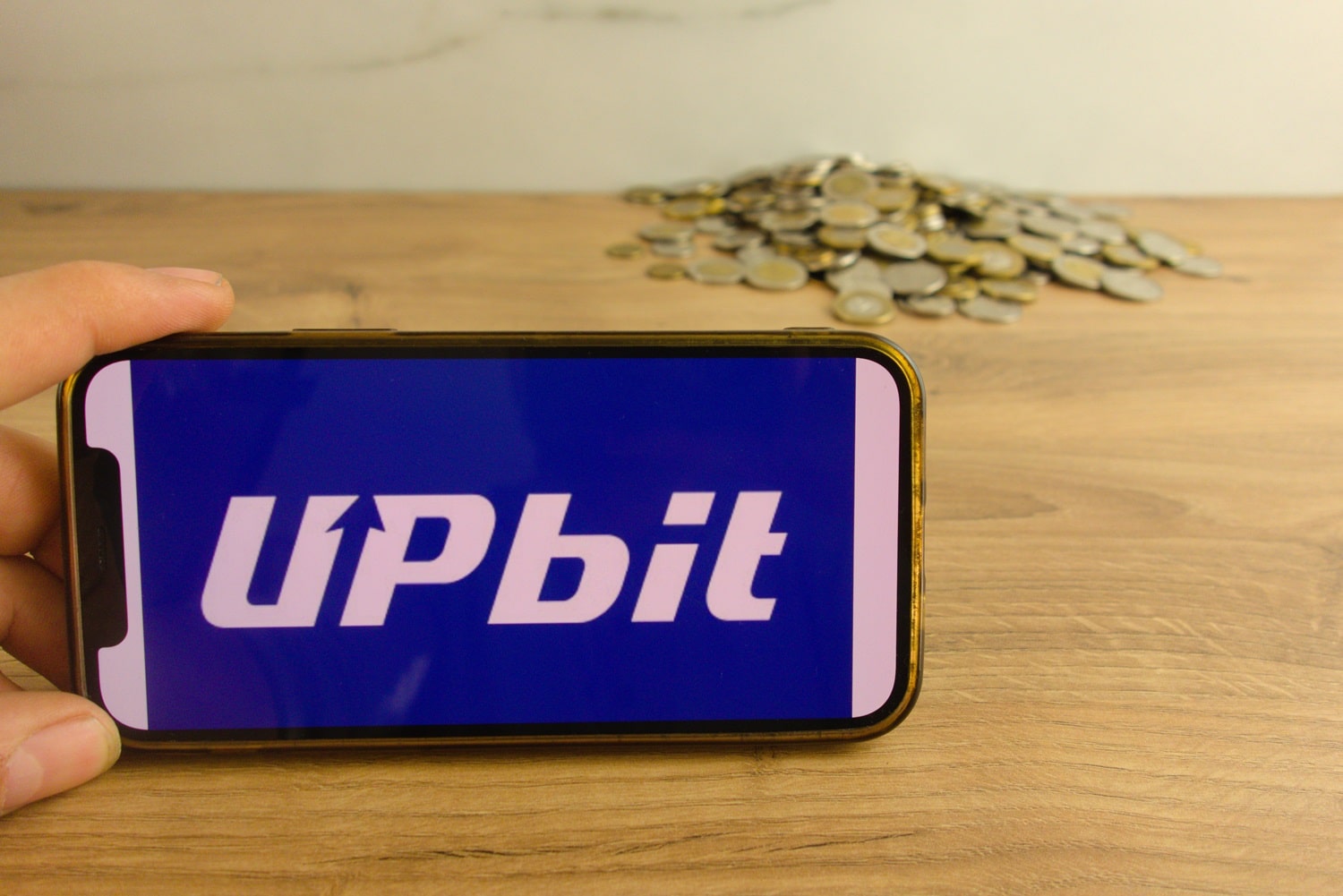 The Upbit cryptocurrency exchange logo on a mobile phone, with a pile of metal coins on a wooden surface in the background.