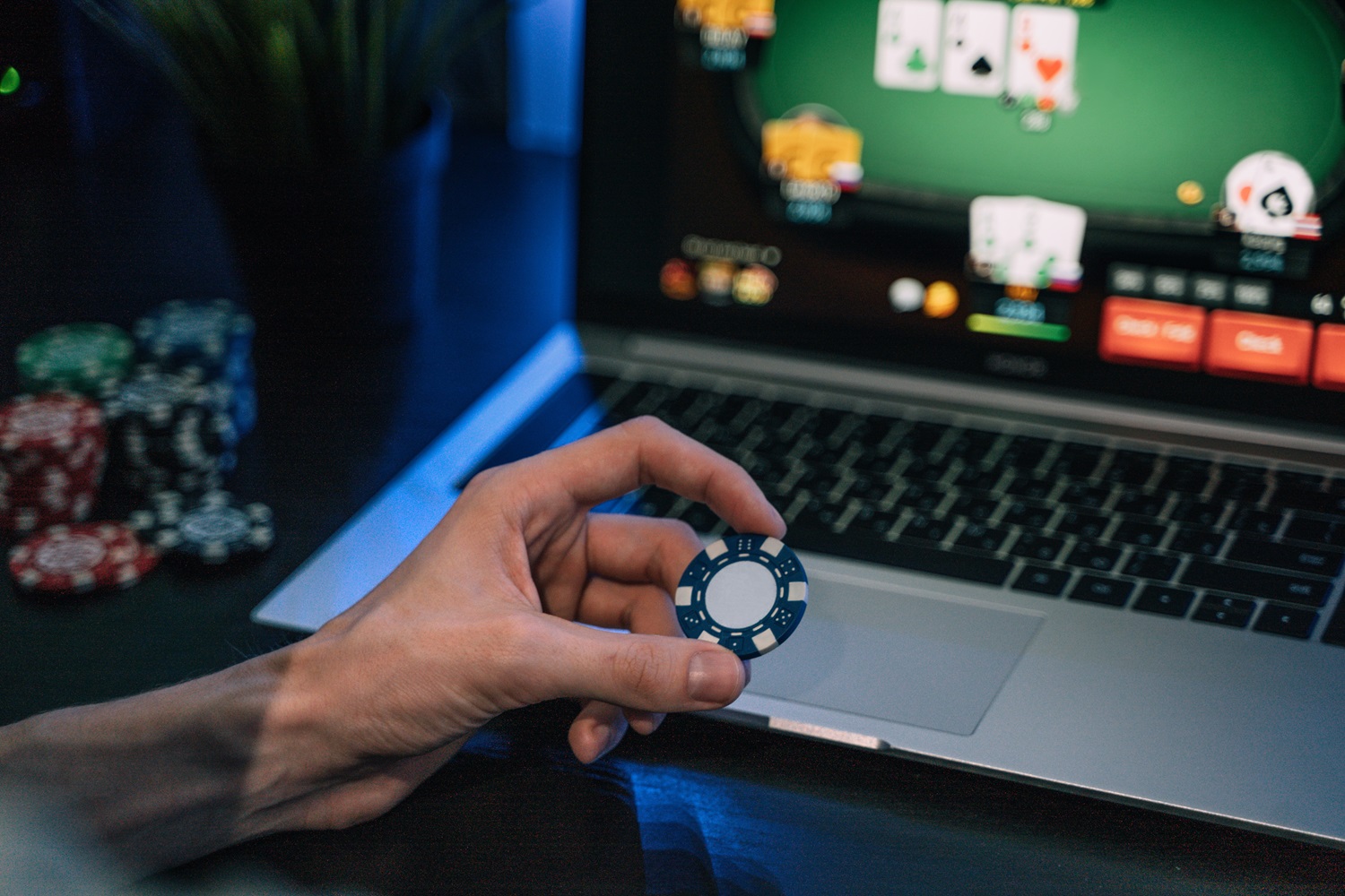 A human hand holds a casino chip in front of a laptop whose screen shows an online poker game.