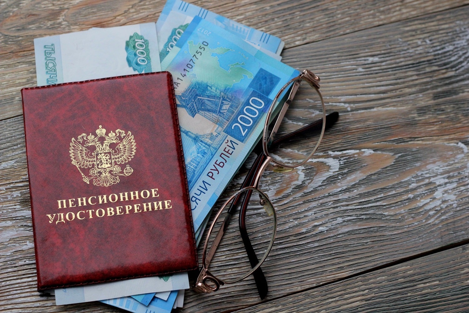Russian banknotes, a Russian pension book, and a pair of glasses on a wooden background.