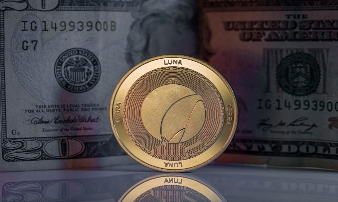 عملة LUNC باللون الذهبيّ ويظهر وراءَها في الخلفية صورةٌ للدولار الأمريكي