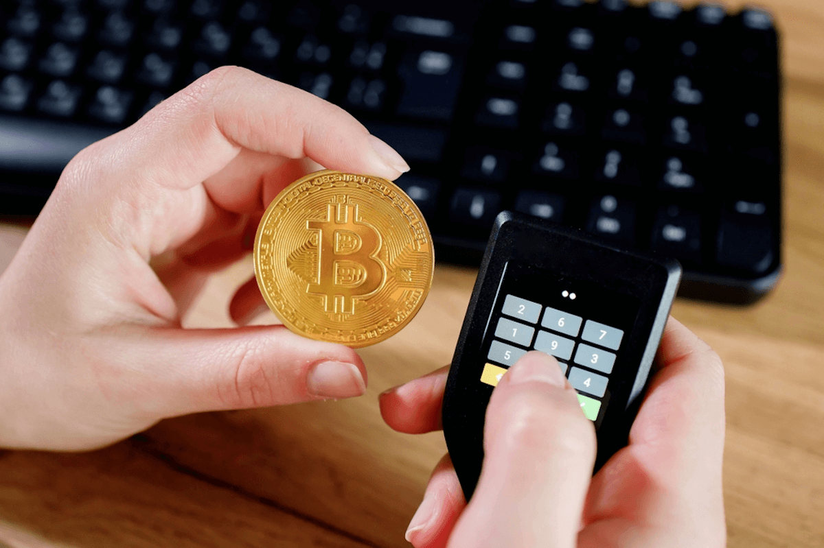 يدٌ تحمل عملةً ذهبيةً عليها شعار بيتكوين والأخرى تحمل جهازاً عليه أرقامٌ وفي الخلفية لوحة مفاتيح