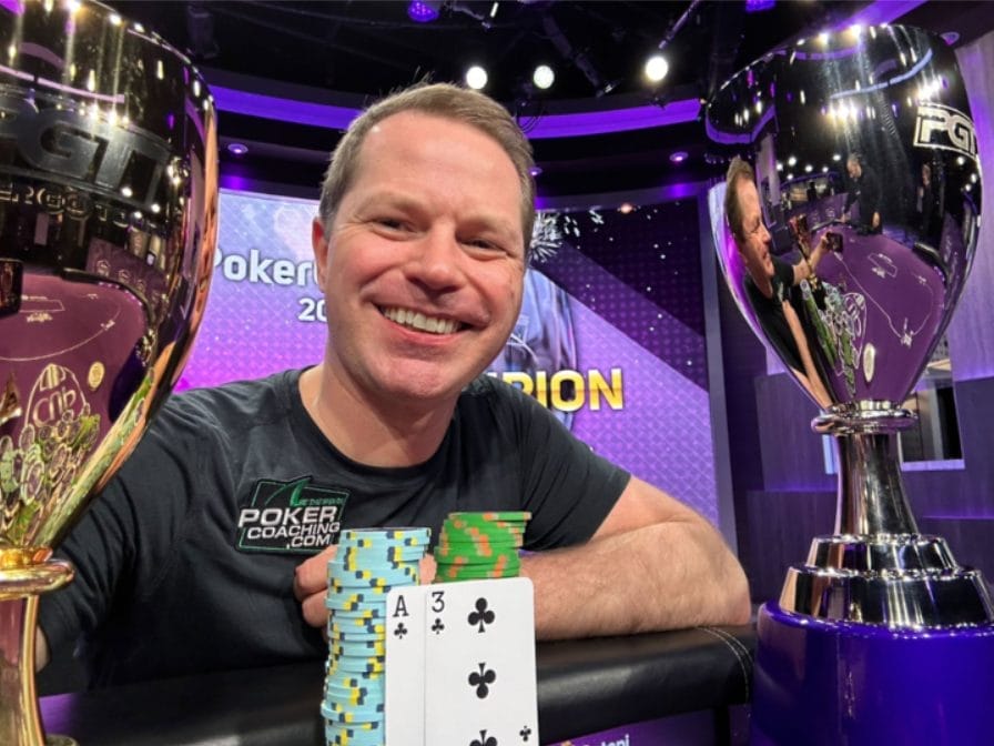 رجل مبتسم يرتدي كنزة عليها شعار موقع PokerCoaching.com وأمامه رقاقات بوكر وورق لعب ويمسك بأحد كأسي بطولة